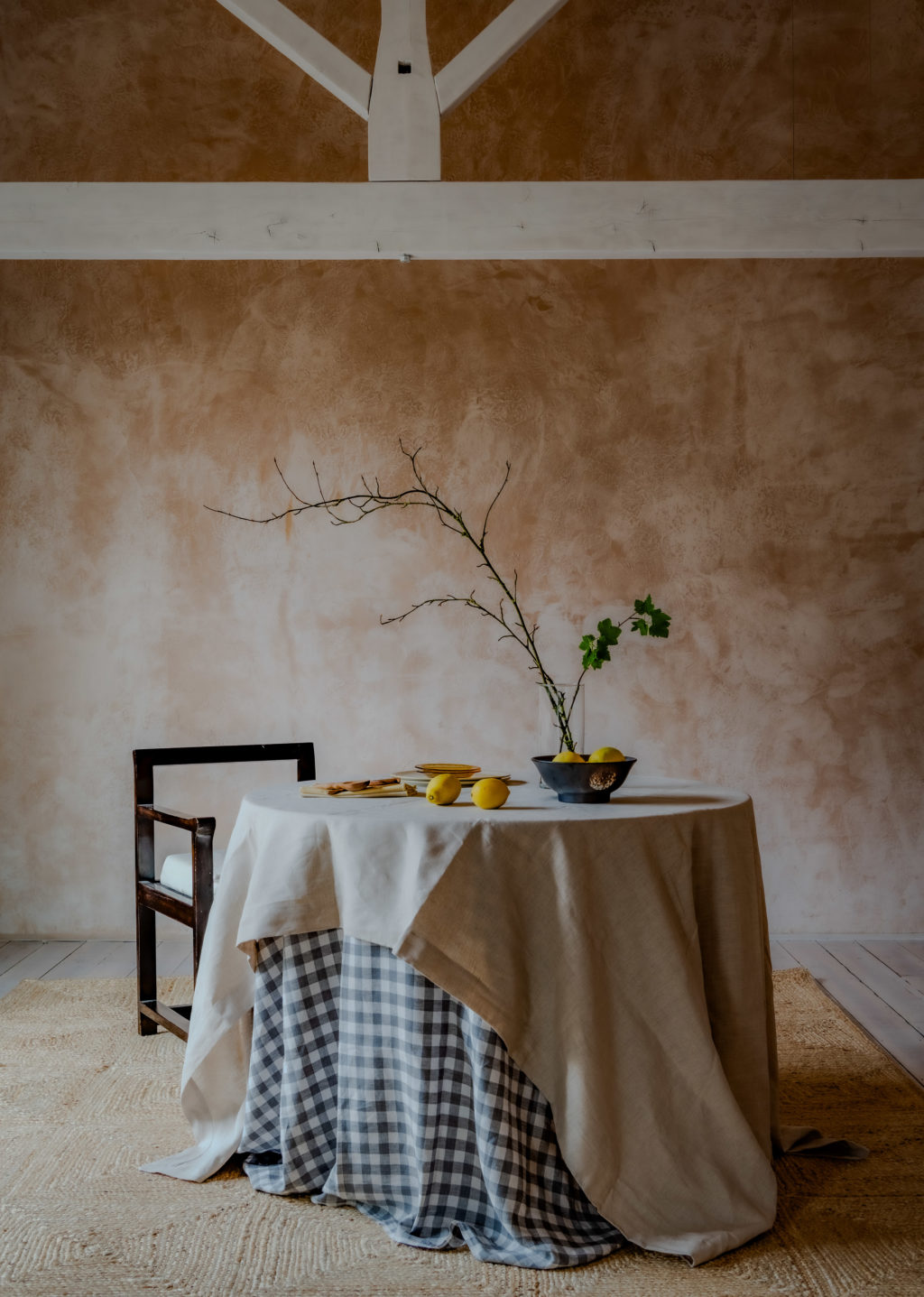 Rustic dining area at The Piggery with textured pink plaster walls, exposed white timber beams and a round table dressed in layered linen tablecloths.