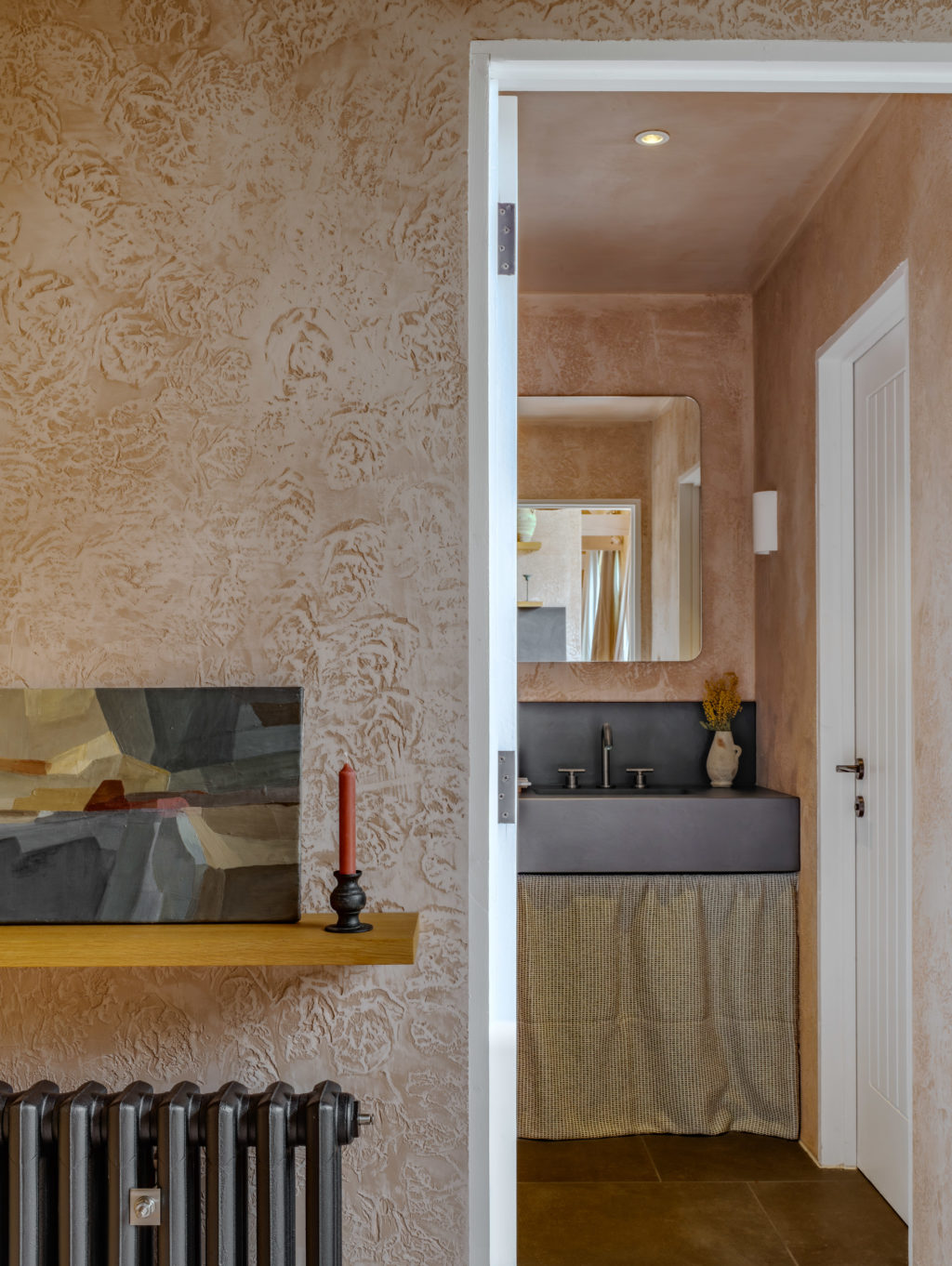 Interior view of The Piggery showing textured pink plaster walls, a timber display shelf and a doorway leading to a bathroom with a black basin and fabric curtain.