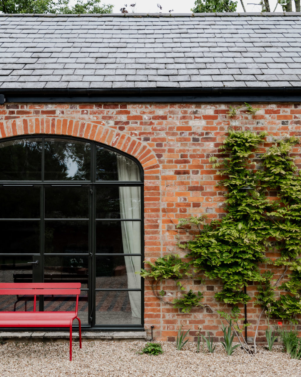 Architectural detail of The Piggery's red brick exterior, featuring a large black-framed arched glazed door, climbing plants and a red metal bench.