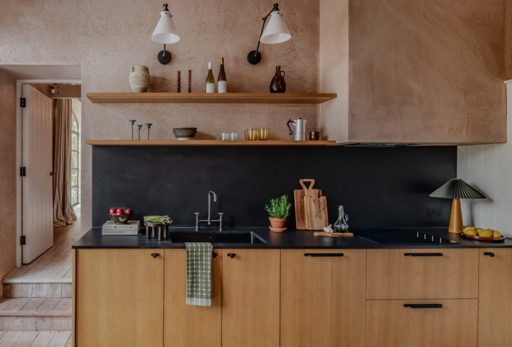 Front view of The Piggery kitchen with Natural Oak Timber Veneer cabinet fronts, black Corston Kilburn handles, Dekton Sirius worktops and oak floating shelves against pink plaster walls.