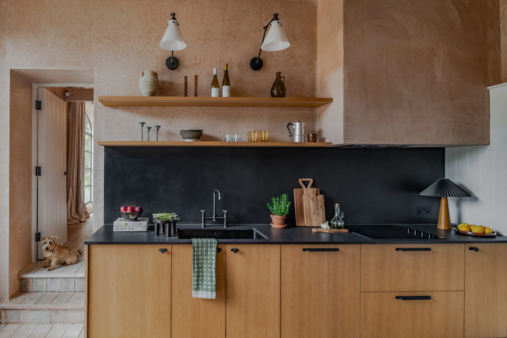 Natural Oak Timber Veneer kitchen at The Piggery with black worktops, open oak shelving and a small dog sitting on the steps beside the cabinetry.