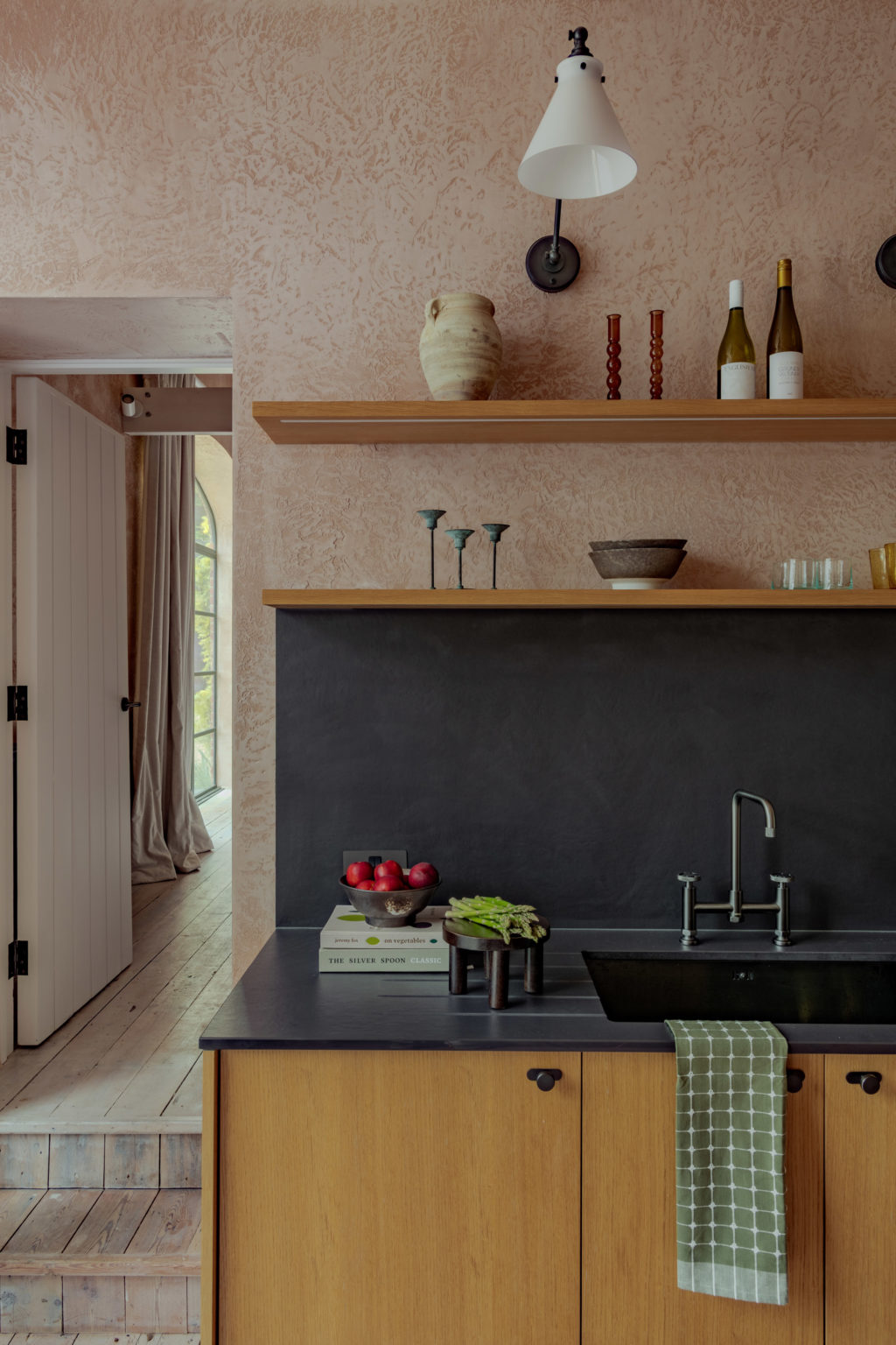 Detail of The Piggery kitchen showing Natural Oak Timber Veneer cabinetry, black sink and worktops, oak floating shelves and decorative ceramics against textured pink plaster.