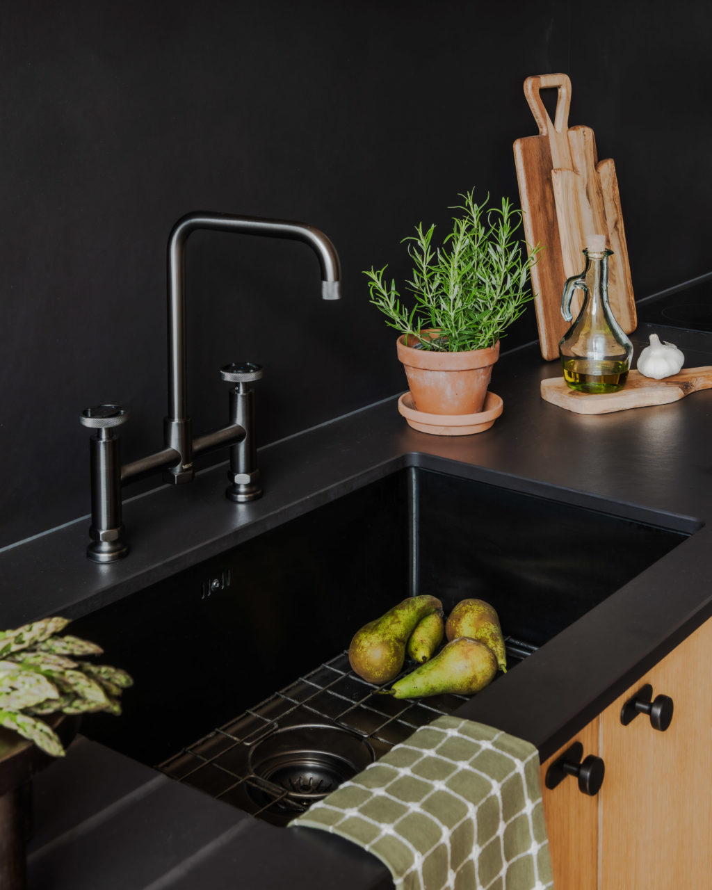 Close-up of the black Abi Interiors Vari sink, Henry Holt bridge mixer tap and Dekton Sirius worktop, styled with fresh herbs, pears and wooden chopping boards.